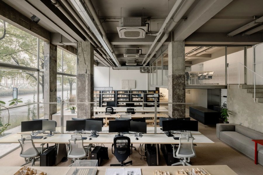 An open-plan office with concrete columns and full-height glazing onto a tree-lined street.