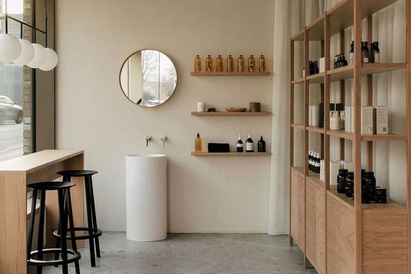 A pale, uncluttered salon interior: a round mirror, a stone basin and open shelves of product.